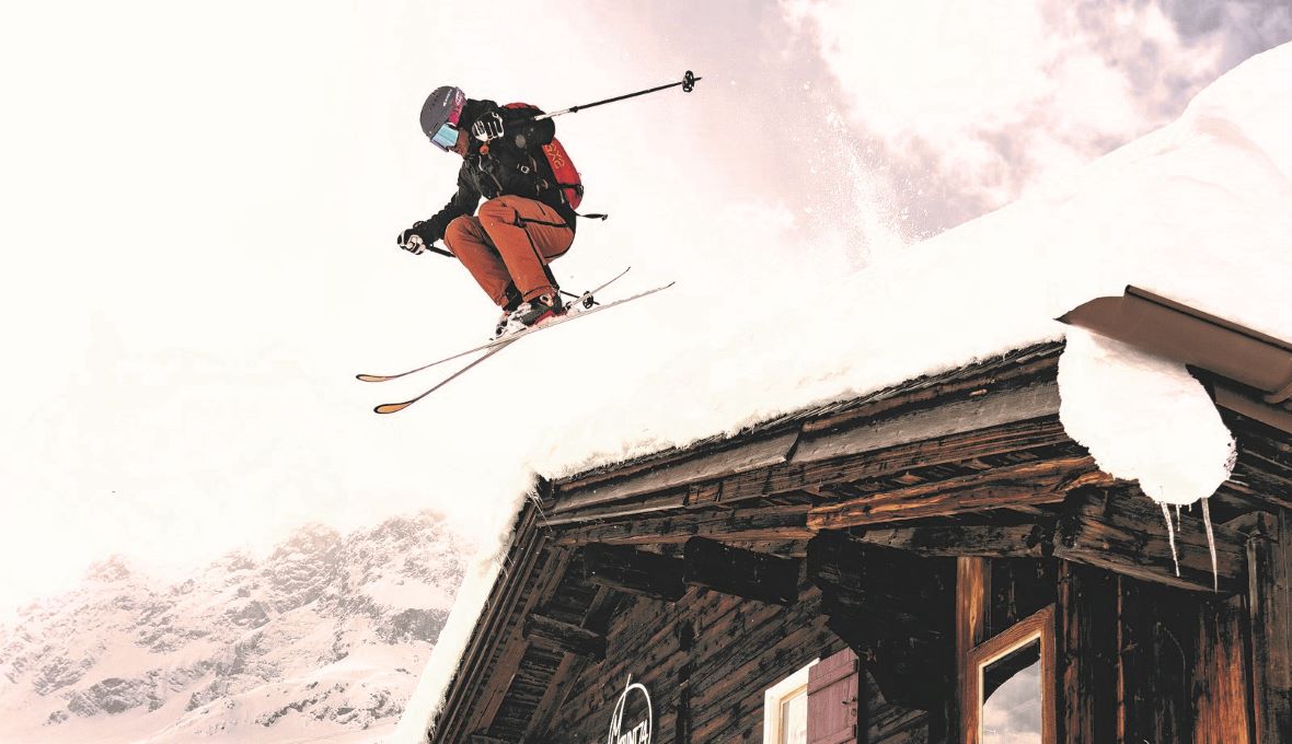 Ein Skifahrer in roter Jacke und orangefarbener Hose führt einen Sprung von einem schneebedeckten Holzdach durch, während er in der Luft schwebt. Der Hintergrund zeigt bewölkte Himmel und Bergspitzen.